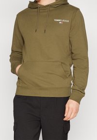 Muž v olivově zelené mikině s kapucí, která má přední kapsu a logo "Tommy Jeans", v kombinaci s černými cargo kalhotami.