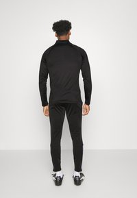 Musta urheilullinen pitkähihainen paita korkealla kauluksella, yhdistettynä samansuuntaisiin mustiin verkkohousuihin. Molemmissa on sileä pinta ja tiukka muotoilu.