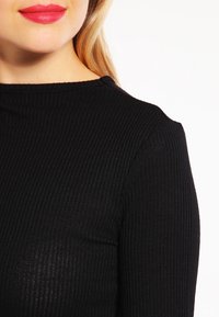 Haut noir côtelé avec un col montant et des manches longues, présentant un tissu texturé et une silhouette ajustée.