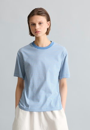 Young person with short hair wearing a light blue and white striped short-sleeve shirt and white pants, standing against a gray background.