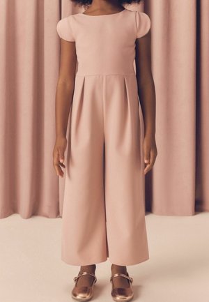 Child stands wearing pale pink jumpsuit with short puff sleeves and pleated wide-leg pants, paired with metallic Mary Jane shoes.