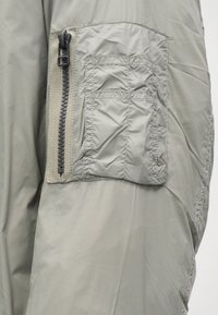 Chaqueta acolchada gris con un bolsillo con cremallera en la manga. La tela es brillante con una textura suave y costuras reforzadas.