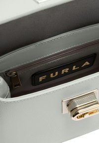 Lichtgrijze leren handtas met een gestructureerd ontwerp. Gouden details, een ritsvak en een stoffen binnenkant met een label waarop "FURLA" staat.