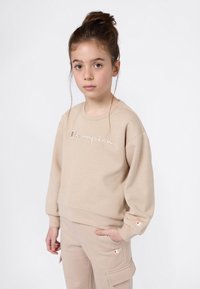 Jeune fille avec les cheveux en chignon, portant un sweat à capuche beige de Champion et un pantalon assorti, debout avec une main dans la poche sur fond blanc.