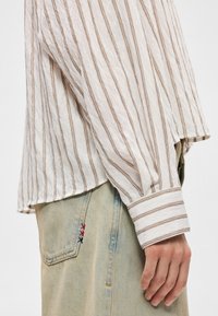 Gros plan d'une main reposant sur le côté, portant une chemise blanche à rayures marron et un jean bleu clair avec de petits détails cousus rouges et bleu foncé.