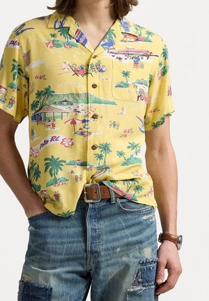 Żółta koszula z krótkim rękawem z tropikalnym nadrukiem plaży i samolotu, noszona z postrzępionymi niebieskimi dżinsami oraz brązowym skórzanym paskiem i zegarkiem.