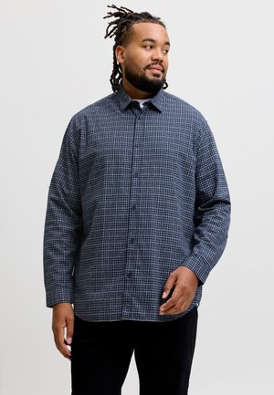 Homme avec des dreadlocks portant une chemise à manches longues bleue et blanche à carreaux, debout devant un fond blanc.