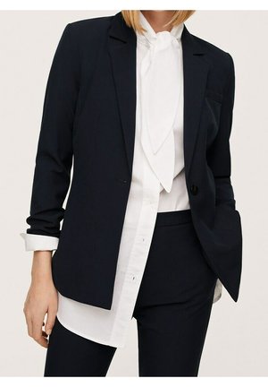 Vrouw draagt een zwart op maat gemaakte blazer en bijpassende broek met een witte blouse met een gebonden nek sjaal.