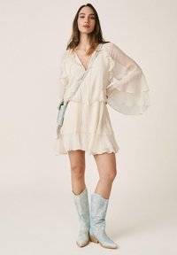 Mujer con un vestido crema con volantes y mangas largas transparentes, botas de piel de serpiente azul y un bolso de hombro azul a juego, posando con una mano en la cadera.