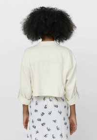 Afgekorte crèmekleurige jas van gestructureerde stof met opgerolde mouwen, gecombineerd met een witte rok met bloemmotief, met blauwe en groene accenten.