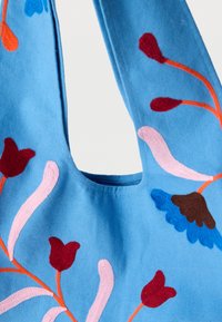 Μπλε βαμβακερή τσάντα με κεντήματα λουλουδιών σε κόκκινο, ροζ και μπλε. Καμπυλωτή κορυφή και υφασμάτινη υφή με λεπτομέρειες.