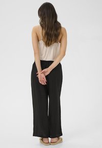 Pantalons larges en lin noir associés à un débardeur beige, vus de dos, avec de longs cheveux ondulés et les mains jointes derrière le corps.