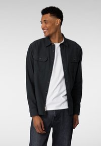 Mørkegrønn skjorte med knapper og to brystlommer, brukt over en hvit skjorte, iført mørkeblå jeans. Glatt stoff, uformell design.