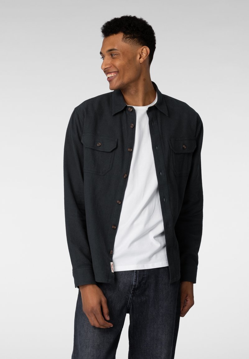 Mørkegrønn skjorte med knapper og to brystlommer, brukt over en hvit skjorte, iført mørkeblå jeans. Glatt stoff, uformell design.