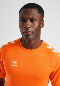 Maillot de sport orange avec un col rond, des manches courtes et des accents en chevrons blancs sur les épaules. Fabriqué en tissu lisse et respirant.