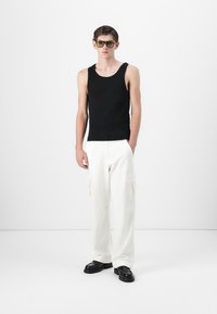 Sort bomulds tanktop, hvide cargo bukser med lommer og sorte sko. Modellen bærer gulfarvede solbriller mod en hvid baggrund.