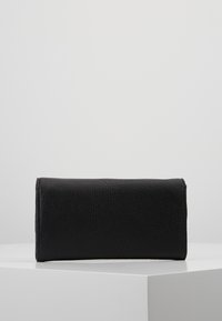 Portafoglio rettangolare nero con texture, in posizione verticale su una superficie bianca contro uno sfondo grigio chiaro.