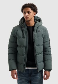 Jongeman in een groene gewatteerde hooded jas over een zwart shirt en blauwe spijkerbroek, staand tegen een egale lichte achtergrond.