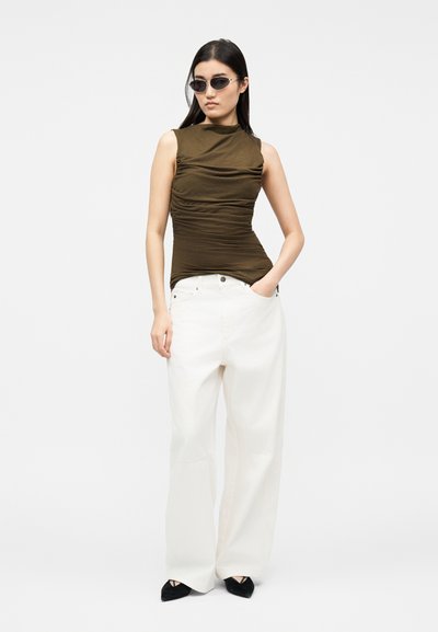 Nő sötét olívazöld, ujjatlan, összehúzott felsőben, fehér bőszárú nadrágban, fekete hegyes cipőben és kis fekete napszemüvegben, kezét zsebre dugva áll.