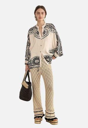 Mujer con camisa oversized beige y negra con estampado, pantalones de crochet color crema, sandalias de plataforma, llevando un bolso negro tejido, de pie sobre fondo blanco.