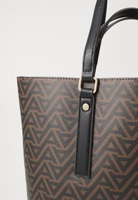 Brun geometriskt mönstrad tygväska med svarta remmar och detaljer i guldmetall. Strukturerad form och texturerat material.