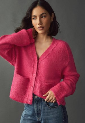 Sart pink cardigan med v-udskæring, to frontlommer og knapper; kombineret med slidte blå jeans. Blød tekstur, løs pasform.
