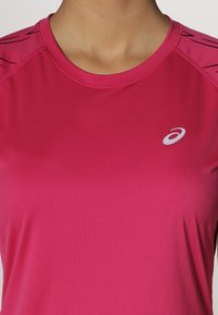 Rosa träningsskjorta i mjukt, lättviktigt tyg. Har en rundad halsringning och kontrasterande svarta linjer vid axlarna. Vit logotyp visas.