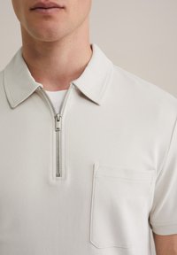 Bela polo majica z zip ovratnikom, kratkimi rokavi in žepom na prsih. Mehka tkanina, gladka tekstura in preprost dizajn.