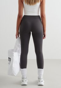 Vrouw in donkergrijze high-waisted leggings, witte sneakers met opgerolde sokken, die een witte totebag vasthoudt en op een lichte vloer staat.