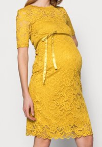 Gul spetsklänning med korta ärmar, blommönster, vågig fåll och satinband i midjan. Lätt, strukturerad tyg.