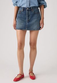 Τζιν μίνι φούστα σε μεσαίο μπλε χρώμα με εφαρμοστή μέση, εμπρόσθιο κούμπωμα με κου μπιά και ραμμένα τελειώματα. Φοριέται με κόκκινα επίπεδα παπούτσια.