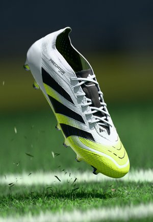 White and neon yellow Adidas Predator soccer cleat with black stripes kicking up grass on a green field near a white line.