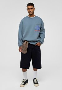 Blauwe sweatshirt met een voork Pocket, met kleurrijke grafische tekst. Zwarte shorts, witte sokken, zwart en bruine sneakers, en een bruine pet.