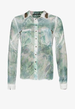 Mujer con una blusa verde translúcida con estampado, vaqueros azules desgastados, botines negros, sosteniendo un bolso de mano negro con una mano en el bolsillo.