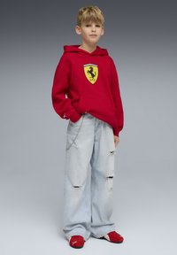 Sweat à capuche rouge avec logo Ferrari, jean bleu clair déchiré oversize, et chaussures rouges. La tenue comprend une poche et un détail en chaîne.