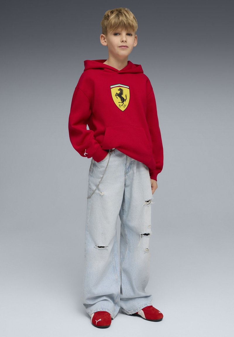 Sweat à capuche rouge avec logo Ferrari, jean bleu clair déchiré oversize, et chaussures rouges. La tenue comprend une poche et un détail en chaîne.