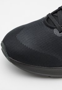 Zapato deportivo negro con parte superior de tela texturizada, que presenta una punta redonda lisa y cordones negros. Patrón sutil en la superficie, suela duradera.