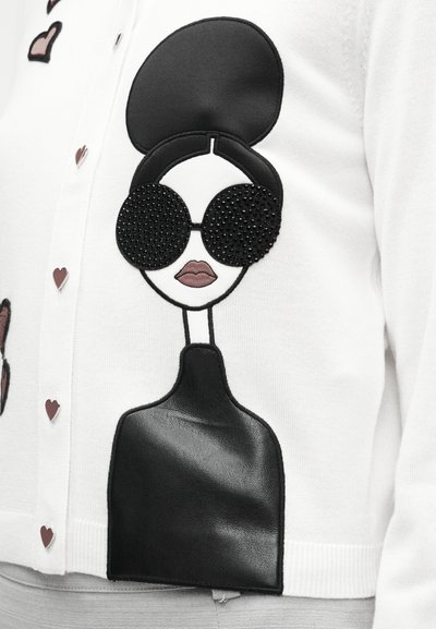 Fehér kardigán, melyen fekete bőr portré látható, hatalmas fekete gyöngyszemüveggel. Apró rózsaszín és piros szív részletekkel díszítve.