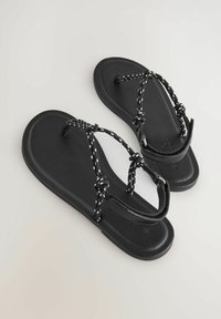 Sandalias planas negras con suela de goma lisa, con tiras trenzadas en negro y blanco para asegurar el pie, diseño minimalista.