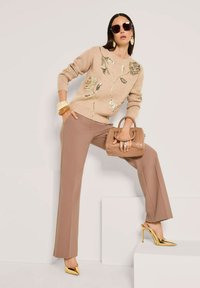 Frau in beigem Pullover mit goldenen Blumenmustern, braunen Hosen, goldenen High Heels, die eine braune Handtasche hält und vor einem hellen Hintergrund posiert.