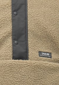 Bézs polár szövet texturált felülettel, sötétszürke patentos betéttel. A fekete címkén a "TAION MOUNTAIN WEAR" felirat olvasható.