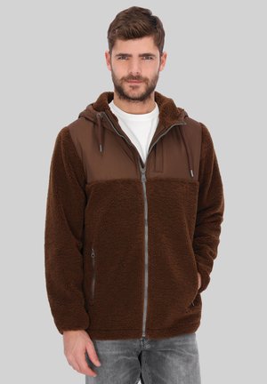 Mann mit Bart, der eine braune Fleecejacke mit Reißverschluss und Kapuze trägt, steht vor einem einfarbigen grauen Hintergrund.