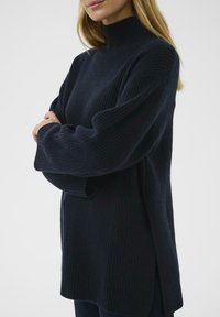 Námořnický pletený svetr s vysokým krkem a uvolněným střihem, s dlouhými rukávy a bočními rozparky pro snadný pohyb.