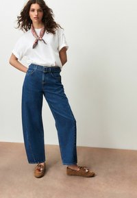 Wijde blauwe spijkerbroek met hoge taille, gecombineerd met een wit shirt met korte mouwen en bruine mocassin-achtige schoenen versierd met accessoires.