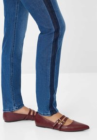 Gambe in jeans blu con dettagli a strisce scure, indossando ballerine bordeaux a punta con doppie fibbie su uno sfondo bianco.