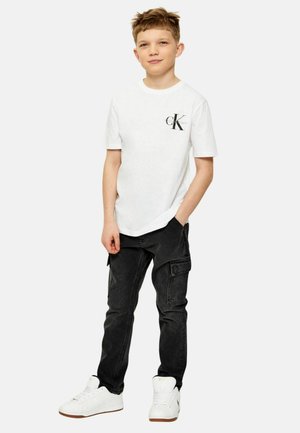 Jongen staat met één hand in zijn zak, draagt een wit Calvin Klein T-shirt, zwarte cargobroek en witte sneakers tegen een effen achtergrond.