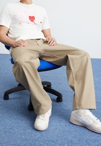 Vit t-shirt med röd grafisk design, beige vida byxor och vita sneakers, sittande på en blå kontorsstol mot en enfärgad bakgrund.