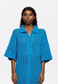 Cardigan blu lavorato a maglia con colletto, caratterizzato da un motivo traforato, maniche corte e chiusura a bottoni. Tessuto testurizzato con vestibilità ampia.