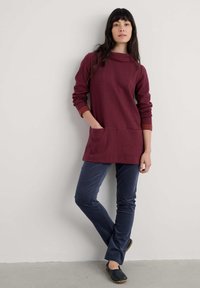 Duge rukave maroon tunike s kapuljačom, s džepom i prugastim detaljima. Uparena s uskim hlačama u mornarsko plavoj boji i crnim klompfama.