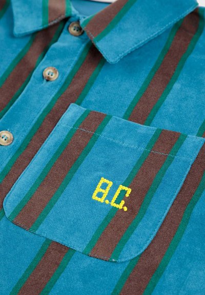 Košulja kratkih rukava u tirkiznoj i smeđoj boji, s vertikalnim prugama. Ima džep na prsima s žutim izvezenim inicijalima "B.C." Dugmad su svijetlo smeđa.
